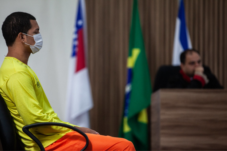 À esquerda, do réu Roberto Marinho Brito (ele veste camisa na cor amarela e calça na cor laranja, está sentado em uma cadeira e usa máscara facial) durante julgamento ocorrido no Fórum de Justiça Ministro Henoch Reis.   Foto: Raphael Alves