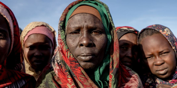 Mulheres e crianças sudanesas deslocadas fazem fila para receber alimentos em um local de distribuição de uma cozinha comunitária da Save the Children no acampamento Daba Naira, em Tawila. Foto: © Unocha/Giles Clarke